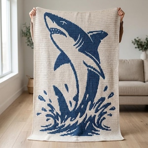 May include: A rectangular crochet blanket featuring a large, dark blue shark leaping out of water against a cream-colored background. The design shows the shark angled upward with splashing water depicted at the base in the same dark blue yarn.