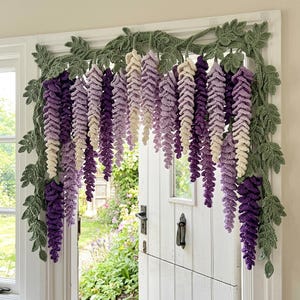 May include: A crocheted wisteria garland in shades of purple, lavender, and cream, with green leaves. The garland is draped over a white door frame, creating a decorative floral accent. The handmade piece adds a touch of nature-inspired charm.