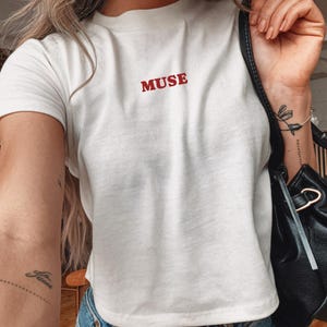 May include: White crew neck t-shirt with the red word "MUSE" printed on the front. The model is 5.2 ft tall and wearing a size S. The shirt is paired with blue jeans and a black purse.