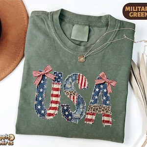May include: Military green t-shirt with the word "USA" appliqued on the front. The letters are made of denim and patterned fabric, with red and white bows. A gold necklace is draped across the shirt.