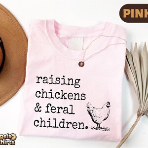 May include: A light pink t-shirt featuring the black printed text 'raising chickens & feral children.' and a graphic of a chicken. A delicate necklace with a small circular pendant rests on the neckline of the shirt.