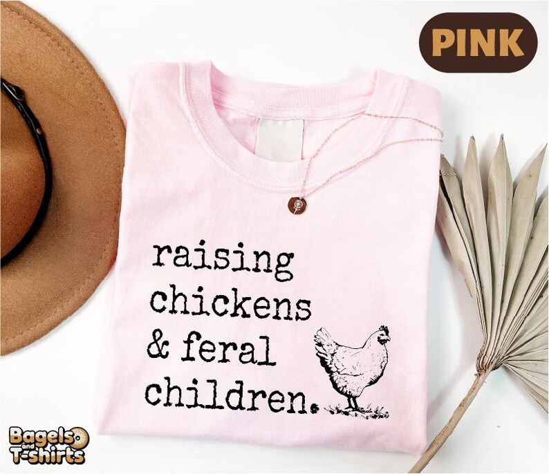 May include: A light pink t-shirt featuring the black printed text 'raising chickens & feral children.' and a graphic of a chicken. A delicate necklace with a small circular pendant rests on the neckline of the shirt.