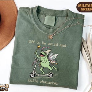 May include: A military green t-shirt featuring a graphic of a frog with fairy wings and a pointed hat riding a scooter while holding a star wand. The text reads, Off to be weird and build character. The shirt has a crew neckline and short sleeves.