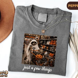 May include: A pepper-colored t-shirt featuring a graphic of a raccoon wearing a ghost costume and holding a coffee cup while pushing a shopping cart filled with Halloween decorations. The text "just a few things" is printed below the graphic.