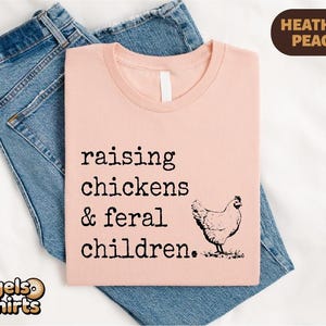May include: A heather peach t-shirt featuring the black text "raising chickens & feral children." and a black line drawing of a chicken. The shirt is layered over a pair of blue denim jeans. A small brown label in the corner reads "HEATHER PEACH".
