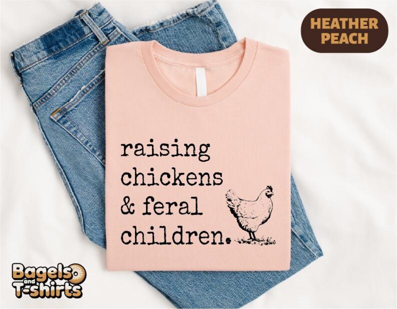 May include: A heather peach t-shirt featuring the black text "raising chickens & feral children." and a black line drawing of a chicken. The shirt is layered over a pair of blue denim jeans. A small brown label in the corner reads "HEATHER PEACH".