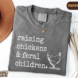 May include: A charcoal gray crew neck t-shirt featuring white screen-printed text that reads "raising chickens & feral children." and a graphic illustration of a chicken. The shirt has a distressed, washed-out appearance and a blank neck label.