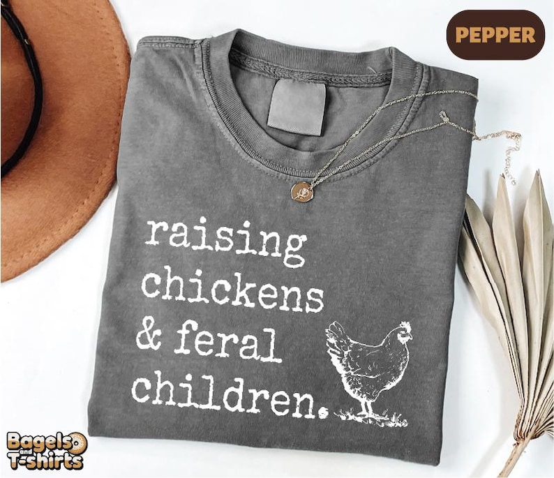 May include: A charcoal gray crew neck t-shirt featuring white screen-printed text that reads "raising chickens & feral children." and a graphic illustration of a chicken. The shirt has a distressed, washed-out appearance and a blank neck label.