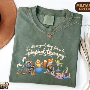 May include: A military green t-shirt featuring a graphic of various animals performing physical therapy exercises. The text reads, it's a good day for physical therapy. The illustration includes a goose, raccoon, duck, fox, frog, and rabbit.