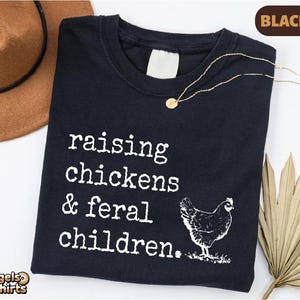May include: A black t-shirt featuring white text that reads "raising chickens & feral children." and a graphic of a chicken. The shirt has a crew neckline and a blank white interior neck label. The design is printed in a typewriter-style font.