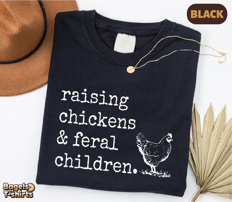 May include: A black t-shirt featuring white text that reads "raising chickens & feral children." and a graphic of a chicken. The shirt has a crew neckline and a blank white interior neck label. The design is printed in a typewriter-style font.