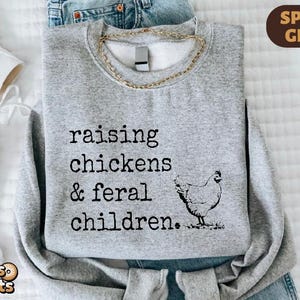 May include: A sport grey crewneck sweatshirt featuring black text that reads raising chickens & feral children. Next to the text is a black line drawing of a chicken. The sweatshirt is layered over blue denim jeans and white sneakers.