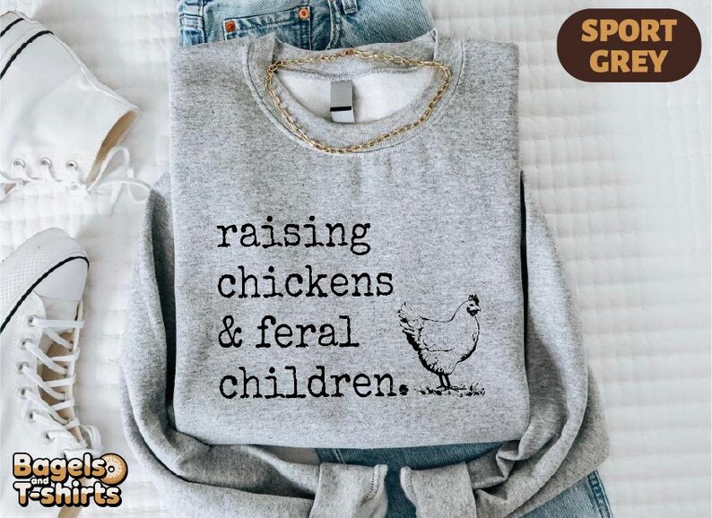 May include: A sport grey crewneck sweatshirt featuring black text that reads raising chickens & feral children. Next to the text is a black line drawing of a chicken. The sweatshirt is layered over blue denim jeans and white sneakers.