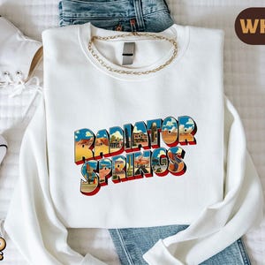 K&ouml;nnte beinhalten: Ein wei&szlig;es Sweatshirt mit Rundhalsausschnitt und dem Schriftzug Radiator Springs in einer fetten, blockartigen Schrift. Die Buchstaben sind mit einer farbenfrohen W&uuml;stenlandschaft gef&uuml;llt, die Felsformationen, blauen Himmel und Geb&auml;ude zeigt.