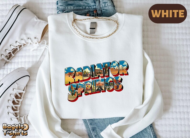 K&ouml;nnte beinhalten: Ein wei&szlig;es Sweatshirt mit Rundhalsausschnitt und dem Schriftzug Radiator Springs in einer fetten, blockartigen Schrift. Die Buchstaben sind mit einer farbenfrohen W&uuml;stenlandschaft gef&uuml;llt, die Felsformationen, blauen Himmel und Geb&auml;ude zeigt.