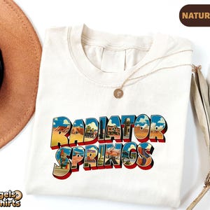 K&ouml;nnte beinhalten: Ein naturfarbenes T-Shirt mit dem Schriftzug Radiator Springs in einer fetten Blockschrift. Die Buchstaben sind mit einer farbenfrohen grafischen Illustration einer W&uuml;stenlandschaft gef&uuml;llt, die Felsformationen und Geb&auml;ude zeigt.
