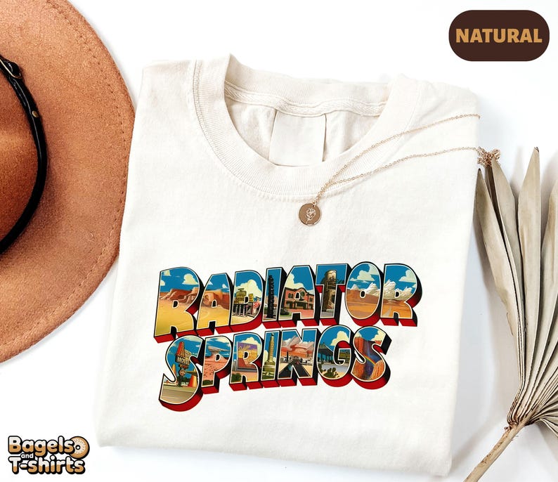 K&ouml;nnte beinhalten: Ein naturfarbenes T-Shirt mit dem Schriftzug Radiator Springs in einer fetten Blockschrift. Die Buchstaben sind mit einer farbenfrohen grafischen Illustration einer W&uuml;stenlandschaft gef&uuml;llt, die Felsformationen und Geb&auml;ude zeigt.