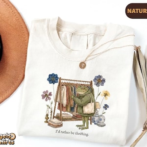 May include: A natural-colored t-shirt featuring a graphic illustration of a green frog wearing a sweater and carrying a tote bag while browsing a clothing rack. The design includes flowers, a lamp, and the text I'd rather be thrifting.