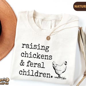 May include: A natural-colored t-shirt featuring the black text "raising chickens & feral children." and a black line-art illustration of a chicken. The shirt has a crew neckline and is displayed with a gold-toned necklace resting on the fabric.