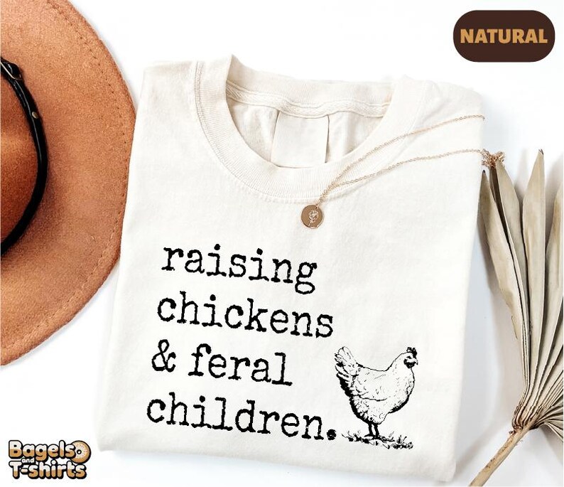 May include: A natural-colored t-shirt featuring the black text "raising chickens & feral children." and a black line-art illustration of a chicken. The shirt has a crew neckline and is displayed with a gold-toned necklace resting on the fabric.
