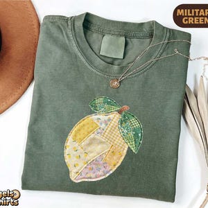 Puede incluir: Camiseta verde militar con un aplique de limón hecho de tela de retazos. El limón tiene patrones amarillos, verdes y florales. Un collar de oro con un pequeño colgante descansa sobre la camisa. La camiseta es de Bagels and T-shirts.