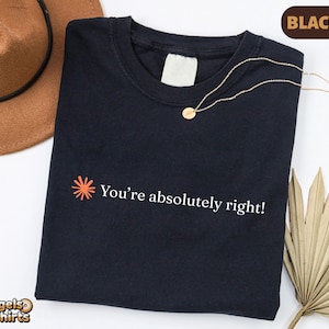 May include: A black t-shirt featuring a small orange starburst icon and the white text "You're absolutely right!" printed on the front. The shirt has a crew neckline and a blank white interior neck label, presented with a brown hat and necklace.