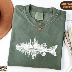 May include: A military green short-sleeve t-shirt featuring a white graphic print of a fish silhouette filled with a forest of pine trees. The shirt has a crew neckline and a blank label area at the inner collar. The text MILITARY GREEN is visible.