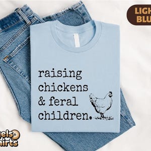 May include: A light blue t-shirt featuring the black text "raising chickens & feral children." and a graphic of a chicken. The shirt is layered over a pair of blue denim jeans. A brown tag in the top right corner displays the text "LIGHT BLUE".