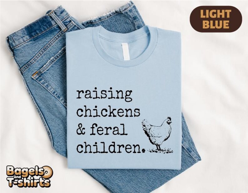 May include: A light blue t-shirt featuring the black text "raising chickens & feral children." and a graphic of a chicken. The shirt is layered over a pair of blue denim jeans. A brown tag in the top right corner displays the text "LIGHT BLUE".
