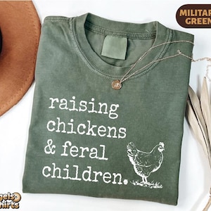 May include: A military green t-shirt featuring the white text "raising chickens & feral children." and a graphic of a chicken. The shirt has a crew neckline and a short-sleeved design, presented with a gold-toned pendant necklace resting on the fabric.
