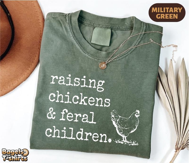 May include: A military green t-shirt featuring the white text "raising chickens & feral children." and a graphic of a chicken. The shirt has a crew neckline and a short-sleeved design, presented with a gold-toned pendant necklace resting on the fabric.