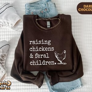 May include: A dark chocolate brown crewneck sweatshirt featuring white text that reads raising chickens & feral children. A white graphic of a chicken is positioned to the right of the text. The sweatshirt is displayed with blue denim jeans.