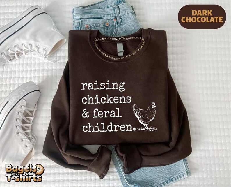May include: A dark chocolate brown crewneck sweatshirt featuring white text that reads raising chickens & feral children. A white graphic of a chicken is positioned to the right of the text. The sweatshirt is displayed with blue denim jeans.