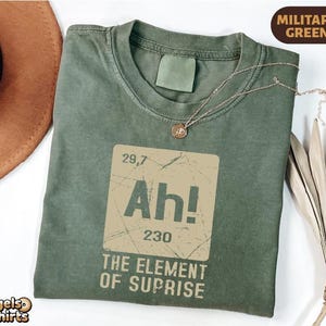 May include: Military green t-shirt with a distressed graphic that reads "Ah! The Element of Suprise." The design resembles a periodic table element. A gold necklace with a pendant is draped across the shirt.