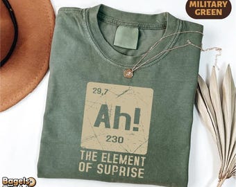 May include: Military green t-shirt with a distressed graphic that reads "Ah! The Element of Suprise." The design resembles a periodic table element. A gold necklace with a pendant is draped across the shirt.