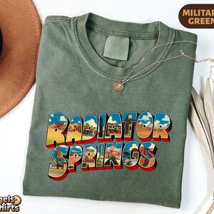 K&ouml;nnte beinhalten: Ein milit&auml;rgr&uuml;nes T-Shirt mit Rundhalsausschnitt und dem Schriftzug Radiator Springs in einer fetten, blockartigen Schrift. Die Buchstaben sind mit farbigen W&uuml;stenlandschaften, Felsformationen und Geb&auml;uden gef&uuml;llt und haben einen roten Schlagschatten.