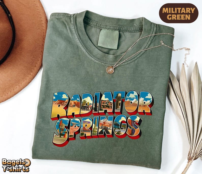 K&ouml;nnte beinhalten: Ein milit&auml;rgr&uuml;nes T-Shirt mit Rundhalsausschnitt und dem Schriftzug Radiator Springs in einer fetten, blockartigen Schrift. Die Buchstaben sind mit farbigen W&uuml;stenlandschaften, Felsformationen und Geb&auml;uden gef&uuml;llt und haben einen roten Schlagschatten.