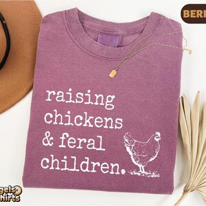 May include: A berry-colored crew neck t-shirt featuring white text that reads raising chickens & feral children. A white graphic of a chicken is positioned below the text. The shirt has a vintage-style wash and a gold-toned necklace rests on it.