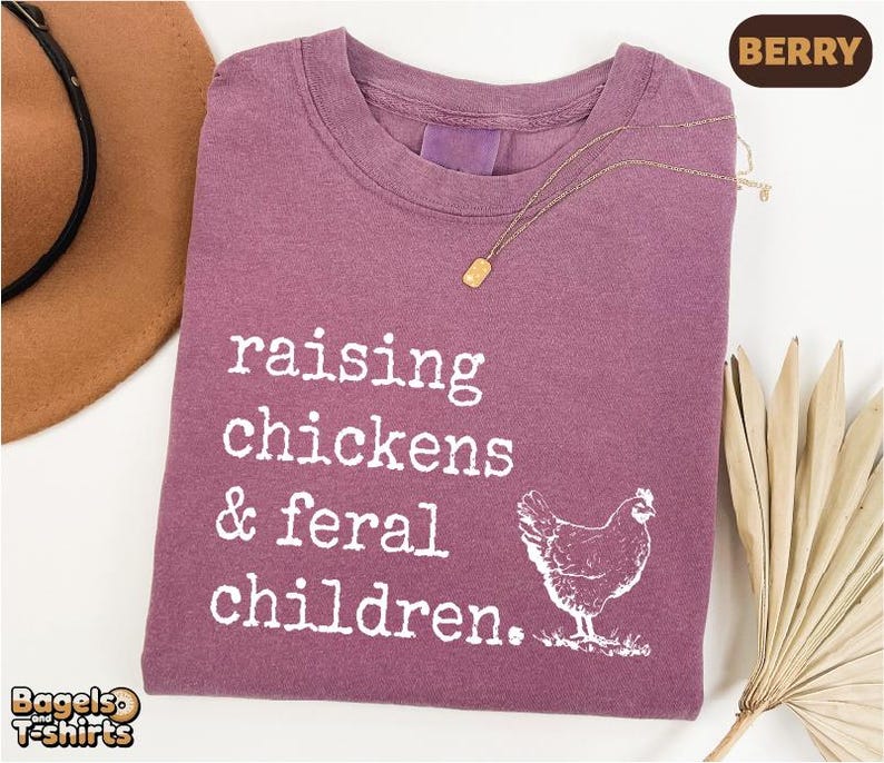May include: A berry-colored crew neck t-shirt featuring white text that reads raising chickens & feral children. A white graphic of a chicken is positioned below the text. The shirt has a vintage-style wash and a gold-toned necklace rests on it.