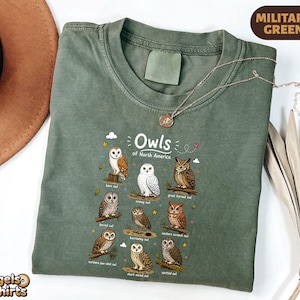 May include: A military green crew neck t-shirt featuring a graphic print titled Owls of North America. The design displays illustrations of nine different owl species with their names printed below each, including barn, snowy, and great horned owls.