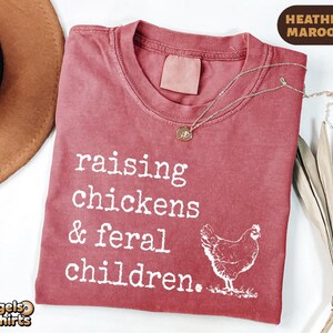 May include: A heather maroon t-shirt featuring white text that reads raising chickens & feral children. A white graphic of a chicken is positioned to the right of the text. The shirt has a crew neckline and a tagless collar design.