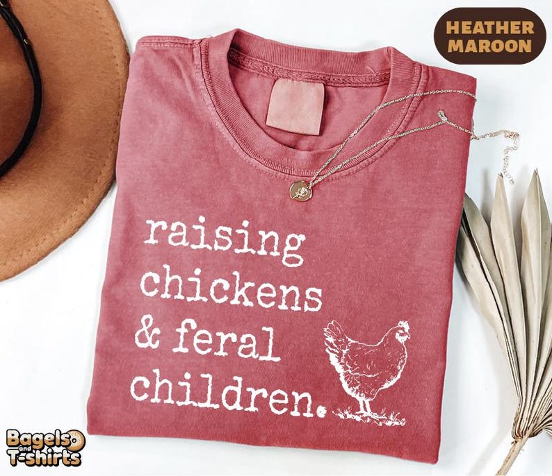 May include: A heather maroon t-shirt featuring white text that reads raising chickens & feral children. A white graphic of a chicken is positioned to the right of the text. The shirt has a crew neckline and a tagless collar design.