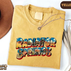 K&ouml;nnte beinhalten: Ein senfgelbes T-Shirt mit Rundhalsausschnitt und dem Schriftzug Radiator Springs in einer fetten, blockartigen Schrift. Die Buchstaben sind mit farbenfrohen W&uuml;stenlandschaften gef&uuml;llt. Eine d&uuml;nne silberne Kette liegt am Kragen.