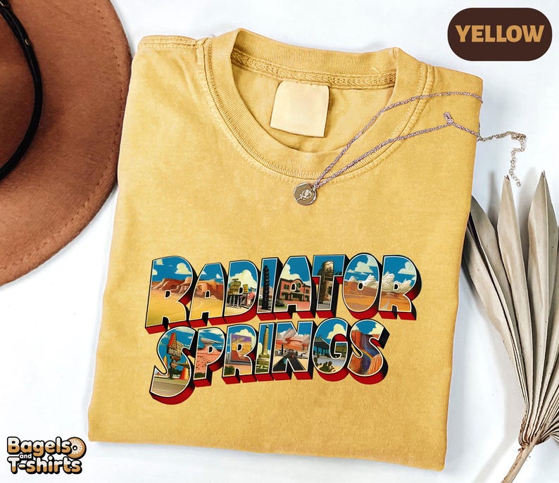 K&ouml;nnte beinhalten: Ein senfgelbes T-Shirt mit Rundhalsausschnitt und dem Schriftzug Radiator Springs in einer fetten, blockartigen Schrift. Die Buchstaben sind mit farbenfrohen W&uuml;stenlandschaften gef&uuml;llt. Eine d&uuml;nne silberne Kette liegt am Kragen.