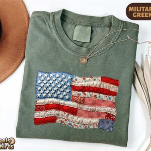 Peut inclure: T-shirt vert militaire avec un drapeau américain en patchwork. Le drapeau présente des tissus bleus, rouges et blancs avec des étoiles et des rayures. Un collier en or avec un pendentif repose sur le t-shirt. Le t-shirt est de Bagels and T-shirts.