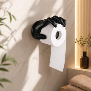 May include: A black hand-shaped toilet roll holder fixed to a wall, holding a roll of white toilet paper. The holder has a matte finish and is designed to resemble a claw. The background is a light-coloured wall with a marble-like pattern.