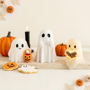 May include: Three white ghost figurines are displayed. One holds an orange jack-o'-lantern bucket, one stands with both middle fingers raised, and one sits while reading a small book. Two cookies shaped like a ghost and a pumpkin are nearby.