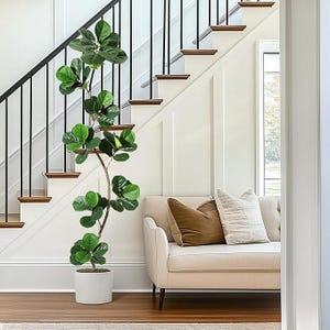 May include: A tall, artificial fiddle leaf fig tree in a white pot. The tree has large, green leaves and a slender, brown trunk. It is placed next to a staircase with black railings and wooden steps. A cream-colored sofa with throw pillows is in the background.