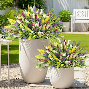 May include: Two large, light gray planters filled with colorful artificial flowers. The flowers are a mix of yellow, purple, pink, and white, creating a vibrant display. The planters are set on a bed of small stones in a garden setting.