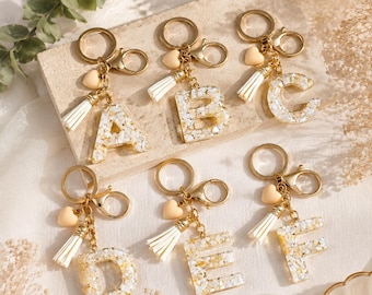 May include: Six resin keychains featuring the letters A, B, C, D, E, and F. Each letter is filled with white and gold flecks, attached to a gold-toned metal keyring and lobster clasp, and accented with a small cream heart charm and a white tassel.